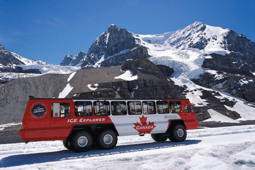 Un autobús rojo y blanco con la leyenda 'ICE EXPLORER' y 'CANADA' estacionado en terreno cubierto de nieve, con montañas al fondo. 1