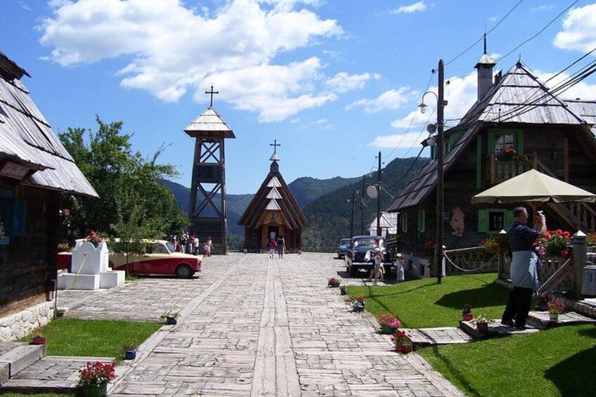 A village street with traditional buildings, a church, and a red car parked on the side. 3