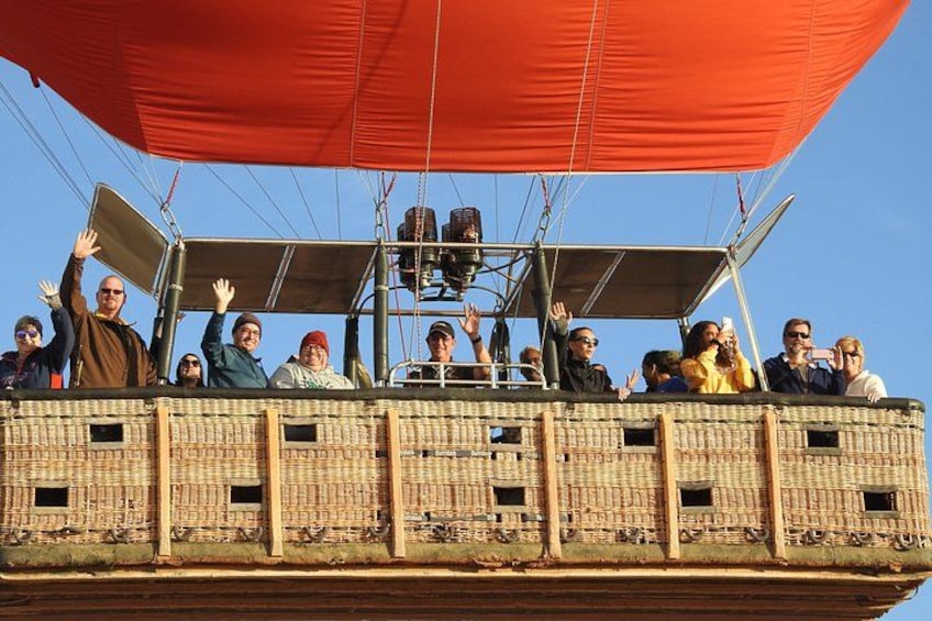 Een luchtballon met passagiers die zwaaien, een mand van wilg en een rode luifel. 3