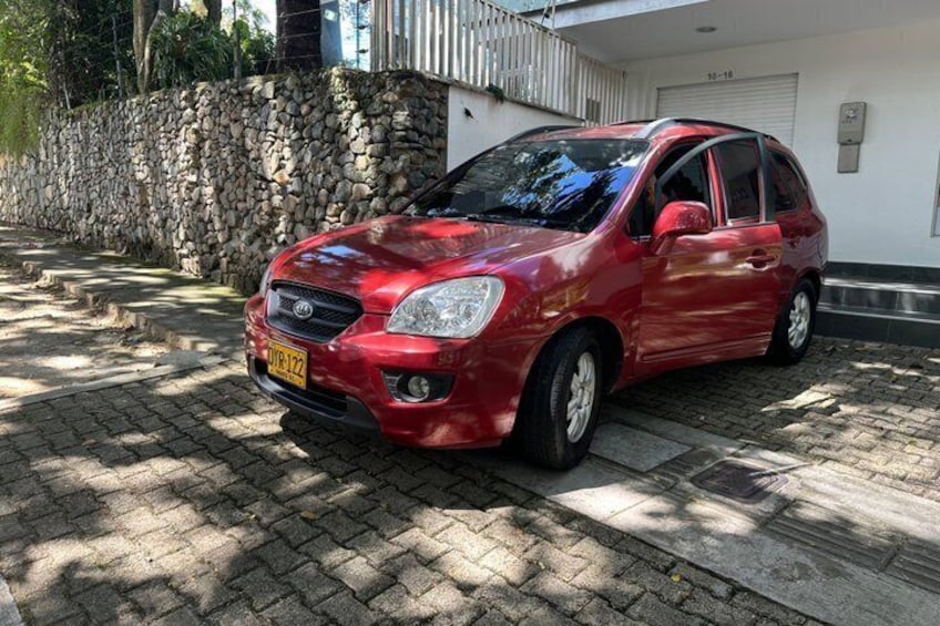 A red car parked on a paved driveway in front of a white building with a stone wall and a metal gate. 3
