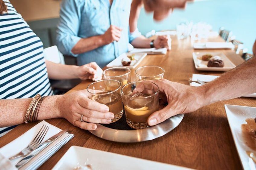 A group of people sitting around a wooden table with glasses of a clear beverage, plates with food, and a tray with more glasses. 3