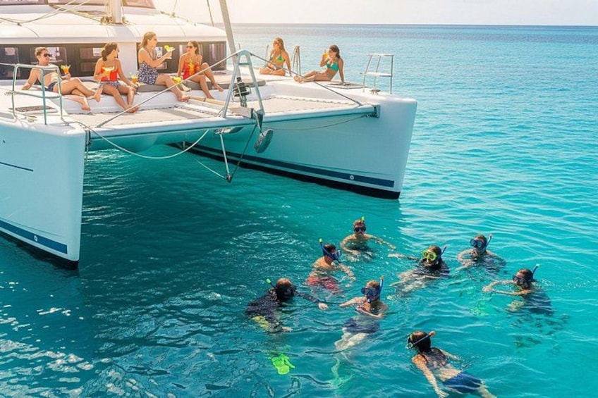 A group of people snorkeling in clear blue water, a white boat with people sitting on the deck, and a calm sea. 3