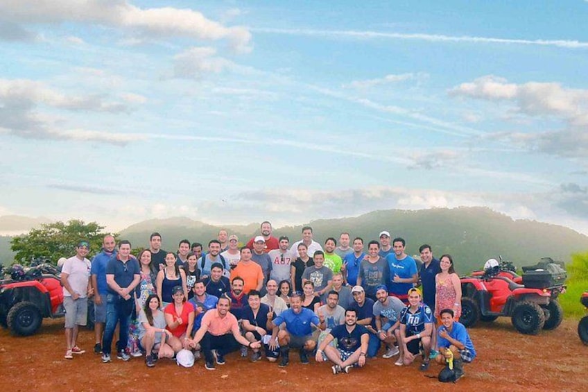 A group of people posing in front of a red vehicle and a scenic landscape with hills. 3
