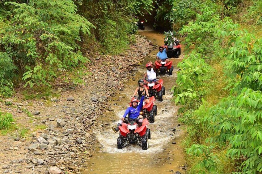 A group of people riding red quad bikes over a rocky stream in a forested area. 3