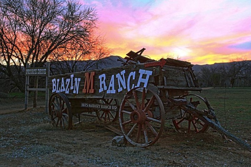 Een houten bord met 'Blazin' M Ranch' en '1875 Mabery Ranch Rd' erop, met een karretje op de achtergrond. 2