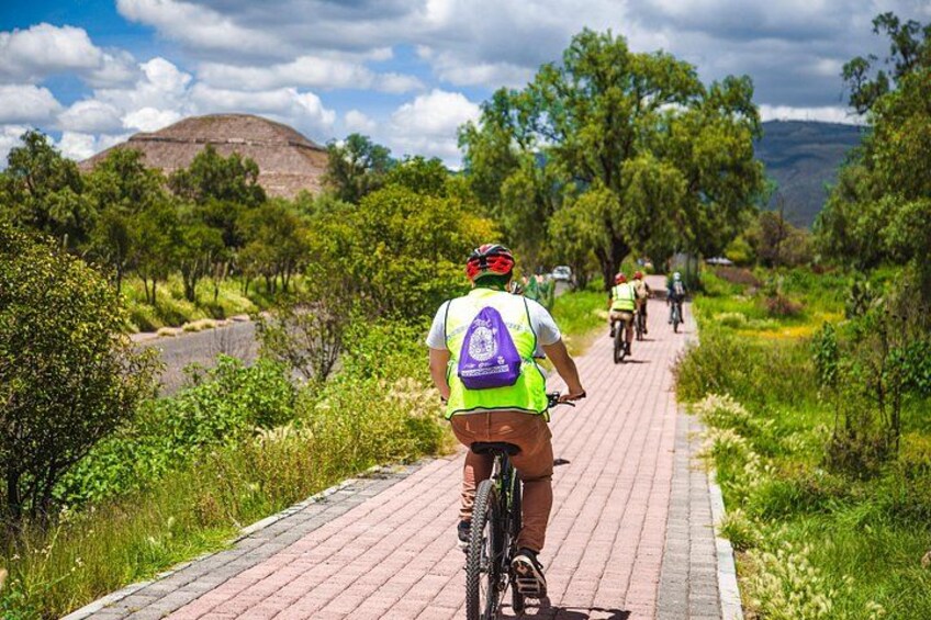 A paved path with cyclists, surrounded by greenery and a hill in the background. 1