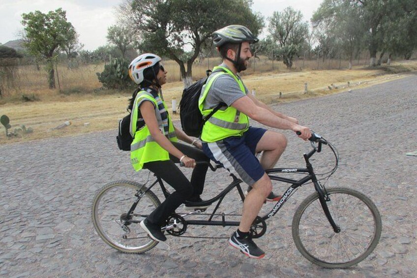 Two people on a bicycle, wearing safety vests, on a cobblestone path with trees and a fence in the background. 3