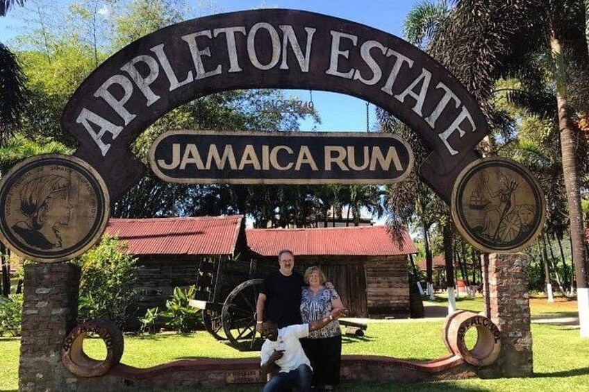 Letrero de Appleton Estate Jamaica Rum con un edificio rústico y una gran rueda de molino al fondo. 1