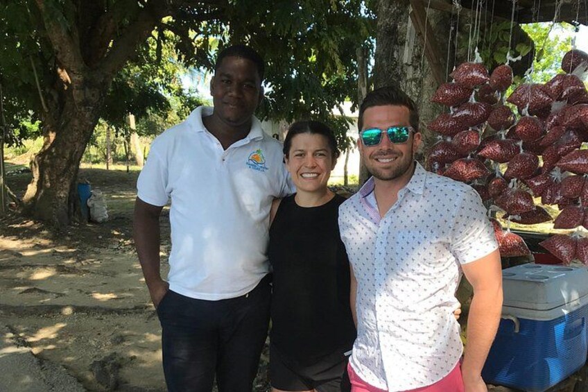 Tres personas de pie bajo un árbol, con una hielera y bolsas de fruta al fondo. 2