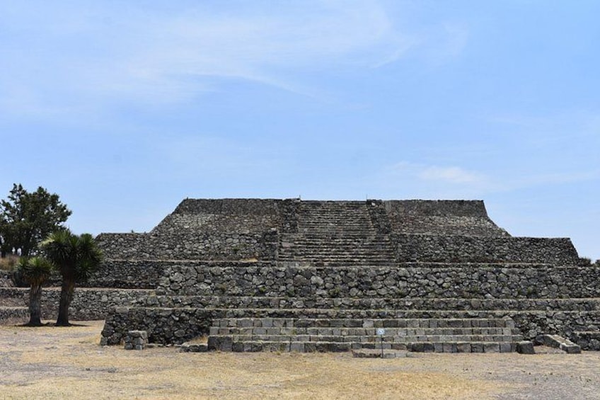 A large stone pyramid structure with multiple levels, surrounded by a dry grassy area with a few trees. 2