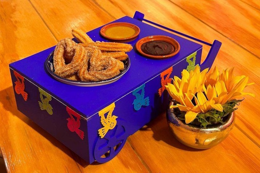 A blue box with colorful designs, containing churros and dipping sauces, and a small pot with yellow flowers. 2