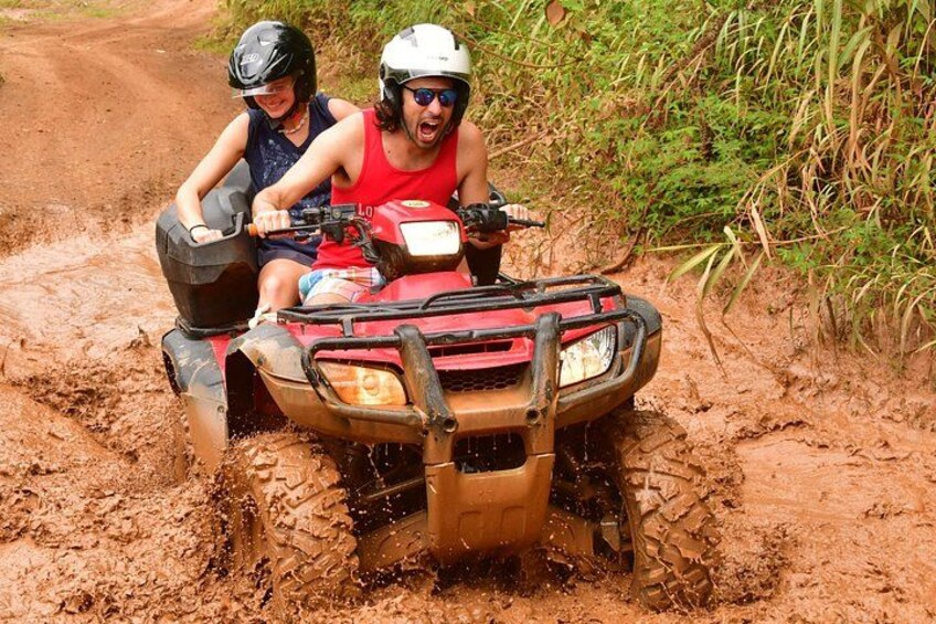 Two people on a red ATV navigating a muddy trail surrounded by vegetation. 5