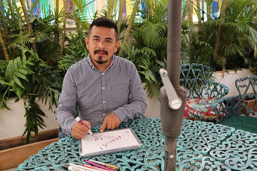 Una persona sentada a una mesa, escribiendo en un cuaderno. Se ven plantas y una silla metálica al fondo. 1