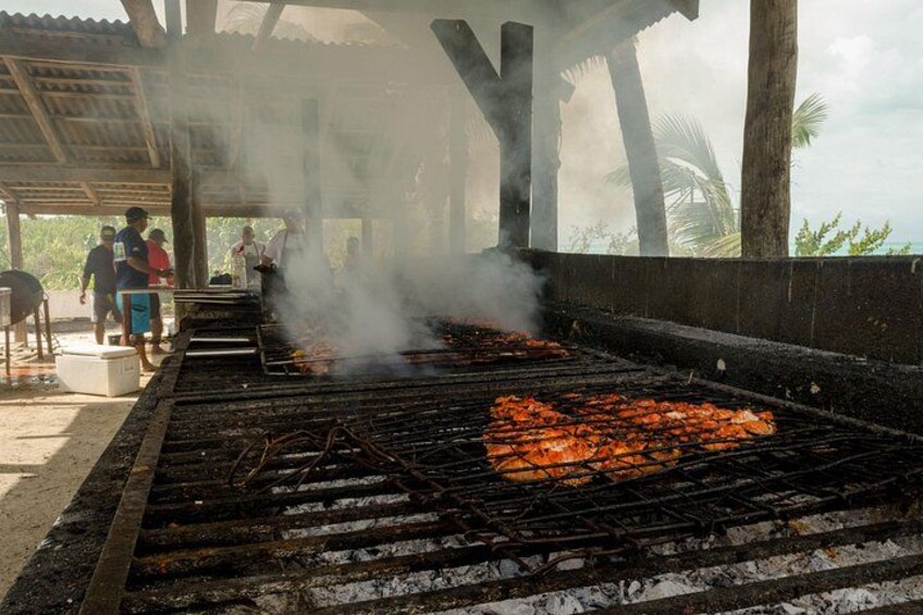 A barbecue grill with food being cooked, people standing around, and a covered area with a view of the ocean. 2