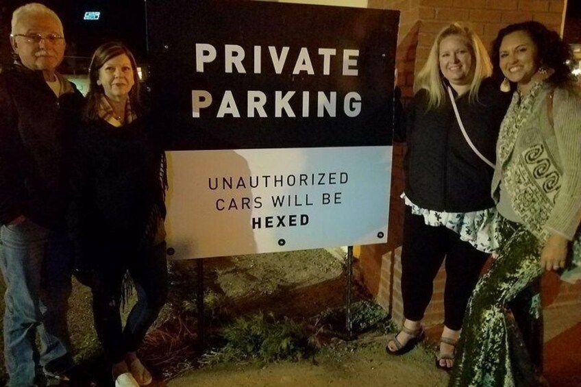 Un grupo de personas de pie frente a un letrero que dice 'PRIVATE PARKING' con una advertencia sobre vehículos no autorizados supuestamente malditos. 4