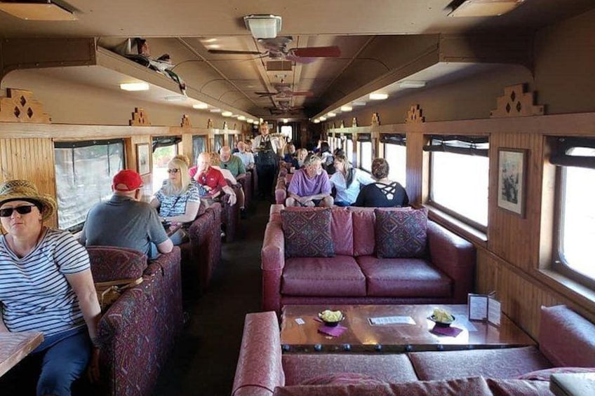 Un vagón de tren con hileras de asientos y mesas, pasajeros sentados y algunos de pie. 2