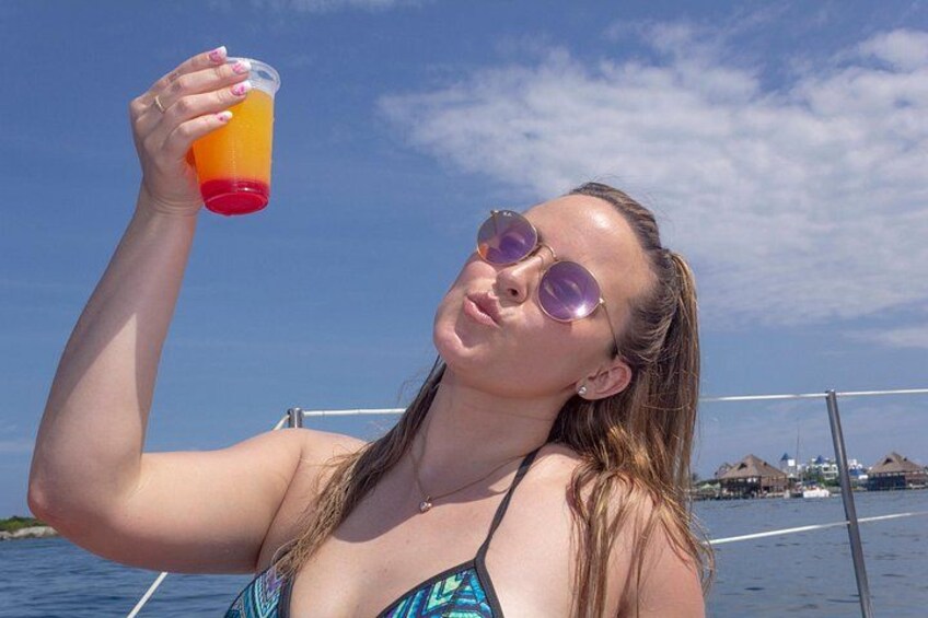 A person holding a glass of orange liquid, wearing a swimsuit, with a clear sky and water in the background. 3