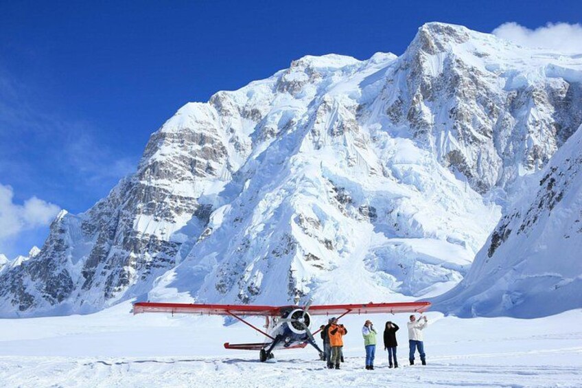 Une montagne enneigée, avec un avion rouge garé dans la neige, et quatre personnes qui se tiennent à proximité. 1