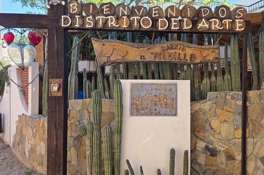 Een houten bord met de tekst 'Bienvenidos Distrito Del Arte' en 'Barrio a Tildillo', omgeven door cactussen. 2