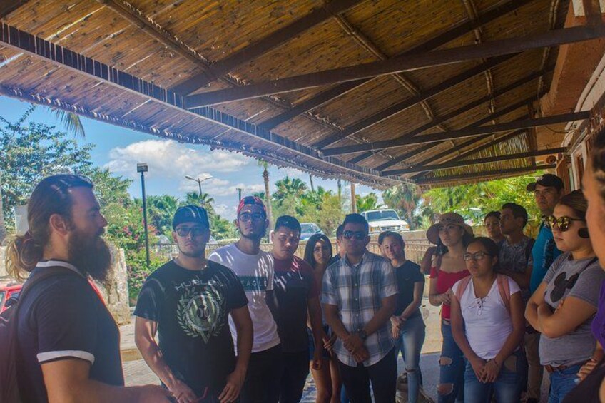 A group of people standing under a wooden shelter with a palm tree in the background. 5