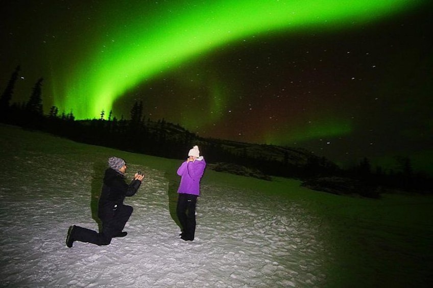 A person kneeling on snow with another person standing, under a green aurora display. 3