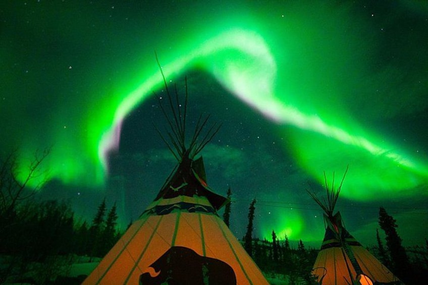 A night scene with a green aurora above teepees in a forest. 1
