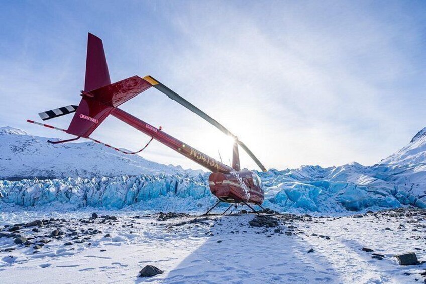 En helikopter med en röd stjärt och gul kaross landar på ett snötäckt område med isformationer och berg i bakgrunden. 3
