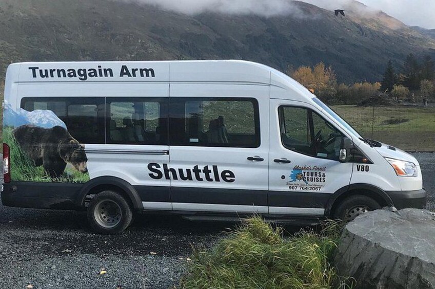 Un autobús lanzadera blanco con la leyenda 'Turnagain Arm' y 'Shuttle', estacionado cerca de una zona rocosa con montañas al fondo. 2