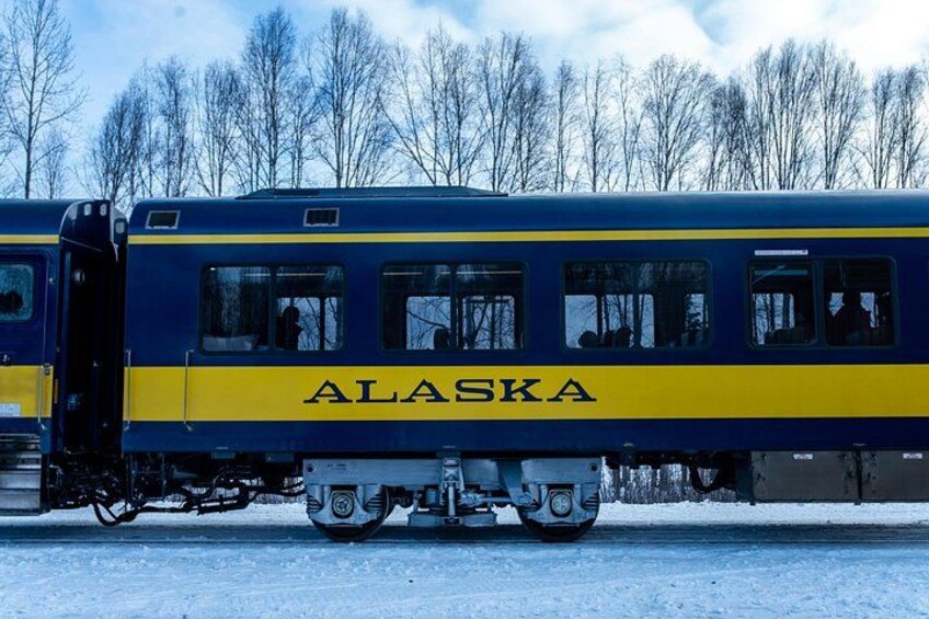 A train with 'ALASKA' written on its side, traveling on tracks with snow on the ground and trees in the background. 4