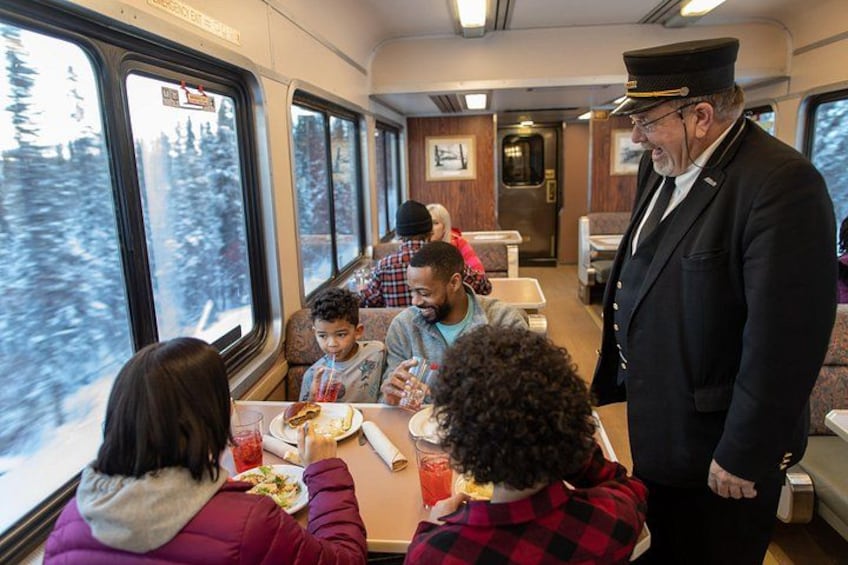 A train dining car with passengers seated at tables, a conductor standing, and a snowy landscape visible through the windows. 3
