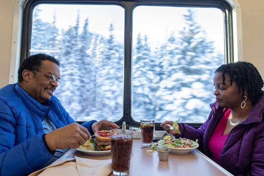 Two people dining inside a vehicle with a snowy forest view. 4