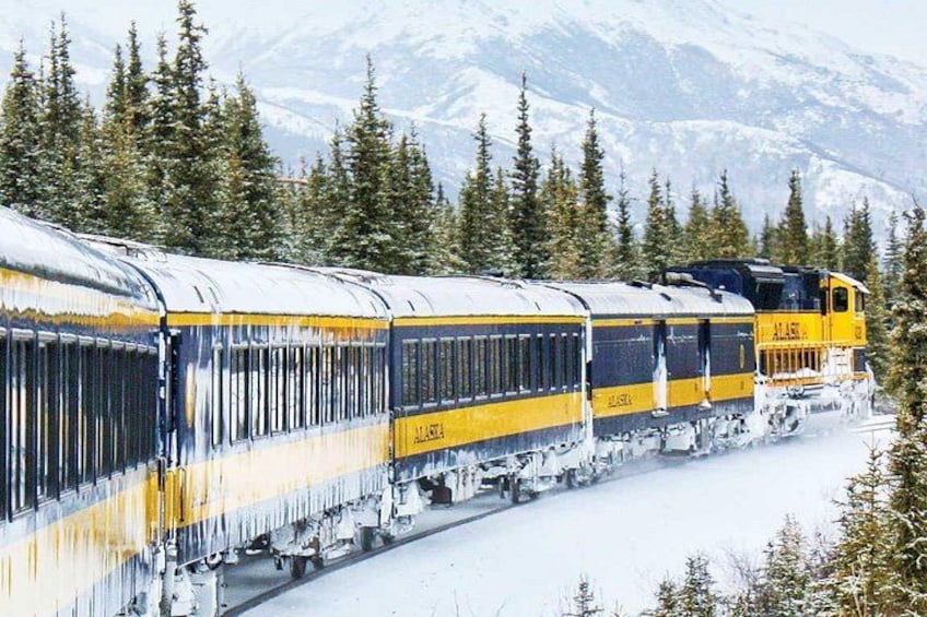 A yellow and blue train with 'ALASKA' written on it, traveling through a snowy forest with mountains in the background. 1