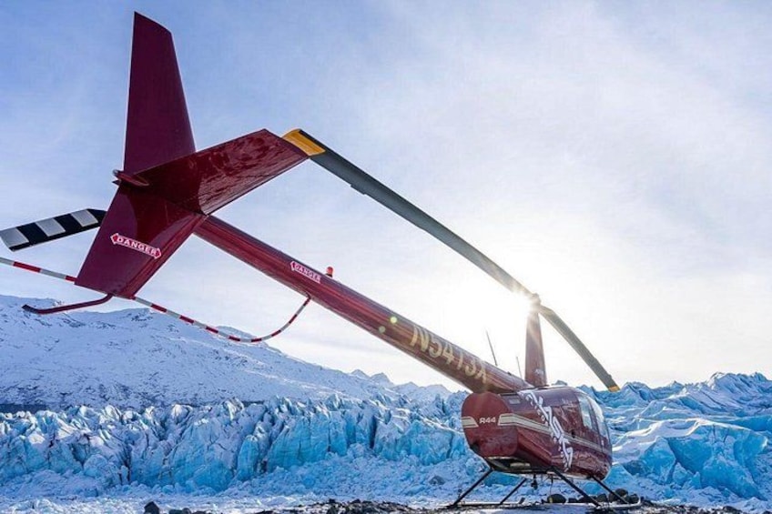 A red helicopter with 'HANSEN' on its tail rotor, parked on a snow-covered glacier with mountains in the background. 5