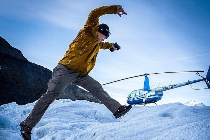 A person in a yellow jacket and brown pants standing on snow with a helicopter in the background. 4