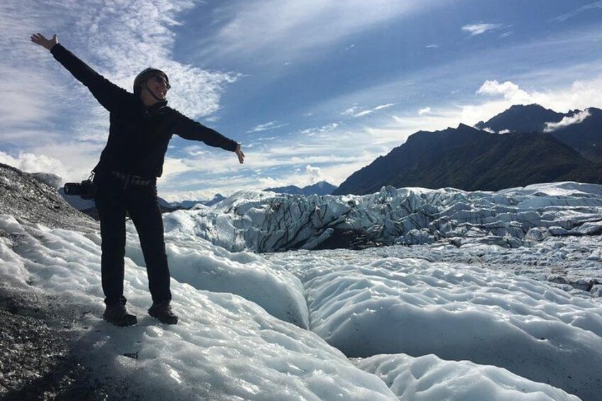 En person står på en glaciär med utsträckta armar, en kamera hänger på axeln och i bakgrunden syns berg. 5