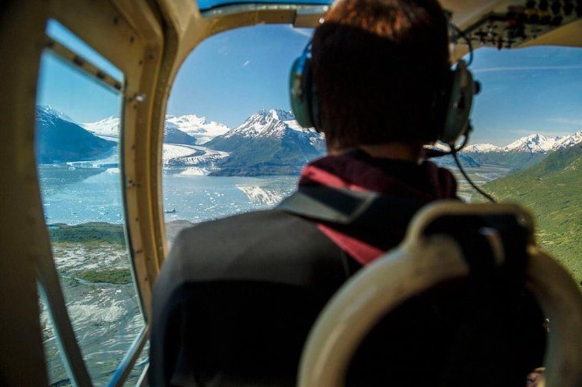 A person in a helicopter cockpit with a view of snow-capped mountains and a glacier below. 5