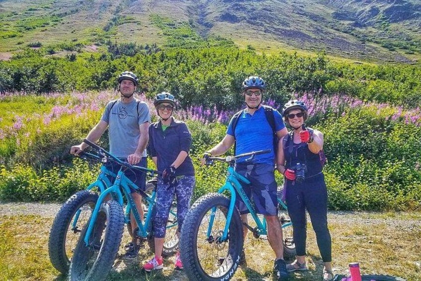 Quatre personnes à vélo tout terrain dans un paysage naturel, avec des collines et des fleurs violettes. 1