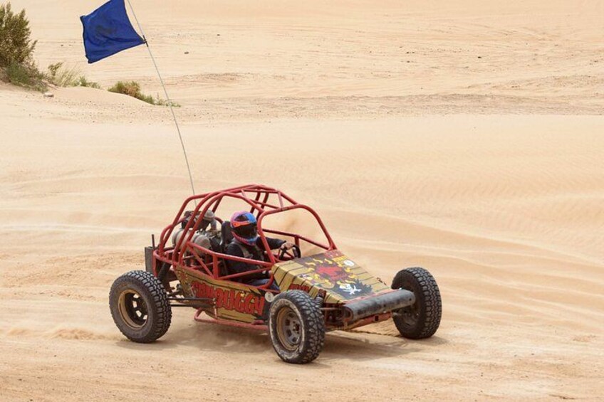 A desert off-road vehicle with a blue flag, traversing sandy terrain. 2