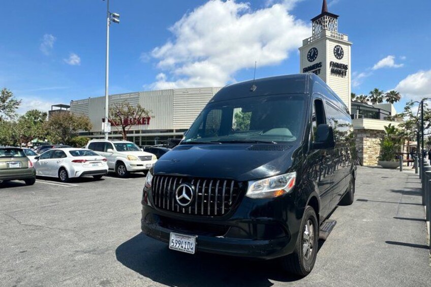 A black Mercedes-Benz van parked in a parking lot with a clock tower and a building labeled 'FARMERS MARKET' in the background. 4