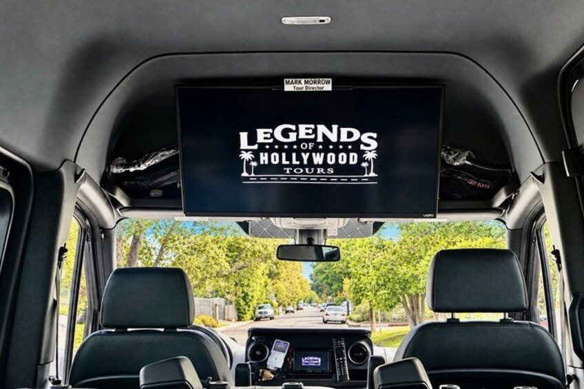 A vehicle interior with a screen displaying 'LEGENDS OF HOLLYWOOD TOURS' and a rearview mirror. 3