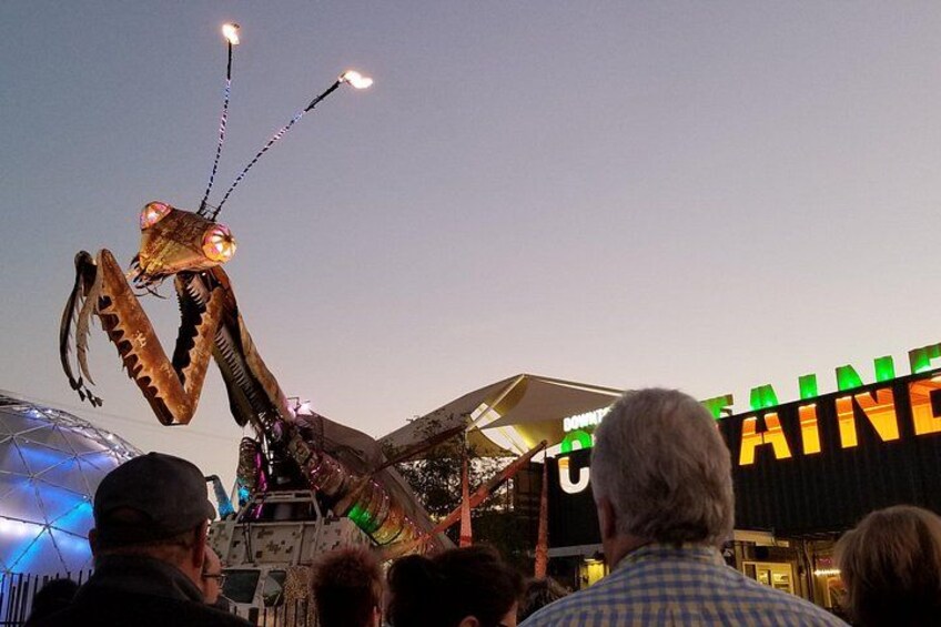 A large insect-like structure with lights, a crowd of people, and a building with illuminated signage. 2