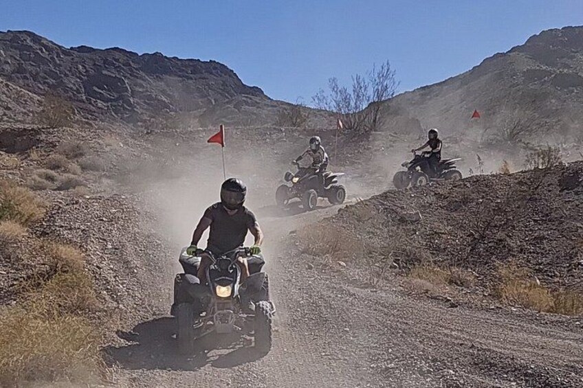 Three riders on quad bikes on a dusty, unpaved road with red flags marking a course, surrounded by dry, rocky terrain. 5