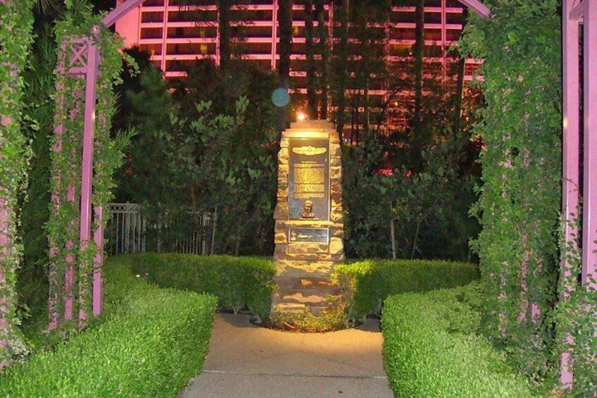 Een stenen monument met inscripties, omgeven door verzorgde hagen en een pad, onder een roze verlichte constructie. 1