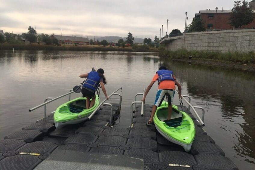 Two people in green kayaks on a dock by a calm river. 4