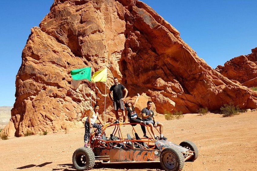Un véhicule tout-terrain avec trois personnes à bord, une grande formation rocheuse rouge et un drapeau en arrière-plan. 2