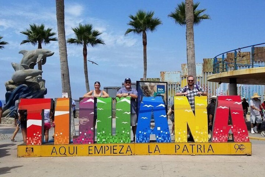 Una playa con palmeras y un gran letrero colorido que dice 'TUNANA', con gente sentada frente a él. 1