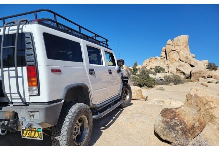 Un Hummer blanc est garé sur une route désertique, avec de gros rochers et un ciel bleu clair en arrière-plan. 2