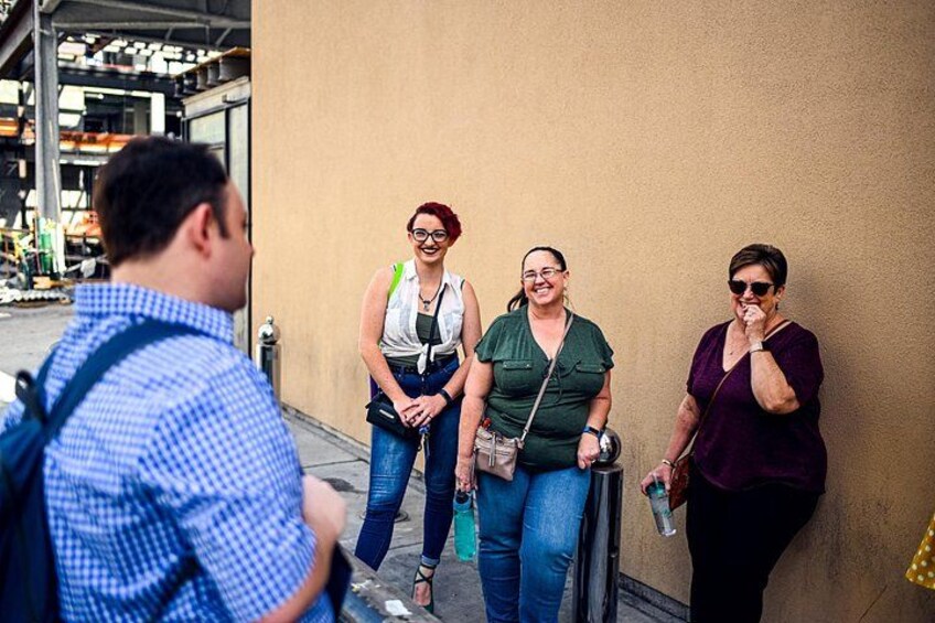 A group of people standing on a sidewalk, a building, and a street with a vehicle in the background. 2