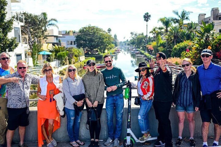 A group of people standing on a bridge overlooking a canal with buildings and palm trees in the background. 1