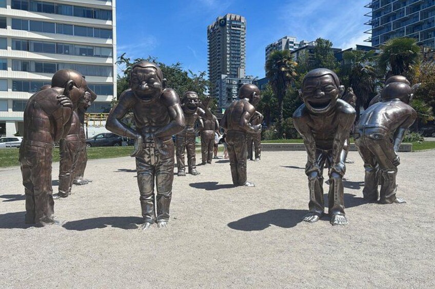 A series of bronze statues of athletes in various poses, set in an open plaza with buildings in the background. 1